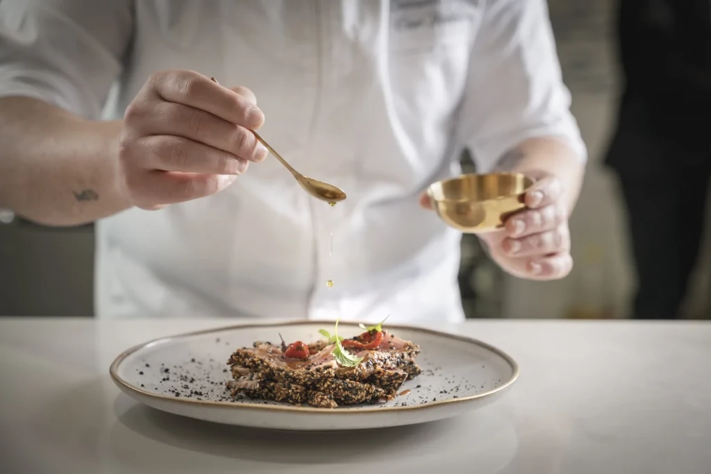 Chef in divisa bianca che irrora con un cucchiaio dorato un piatto gourmet con crosta di semi e guarnizioni rosse